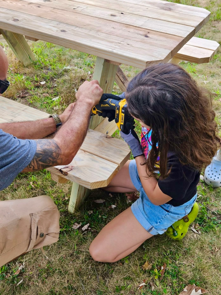 Girl Scout kneels beside a picnic table using a power drill while an adult volunteer assists