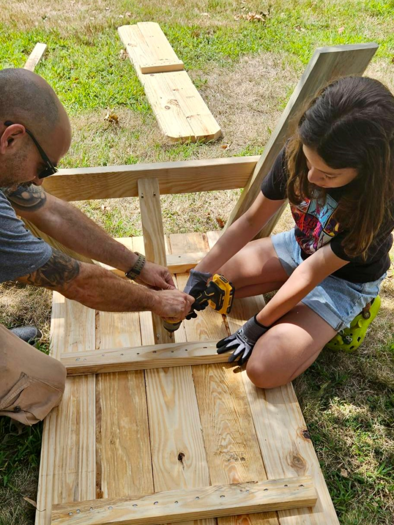 Girl Scout uses a power drill to attach support beams underneath a picnic table at Girl Scout Park