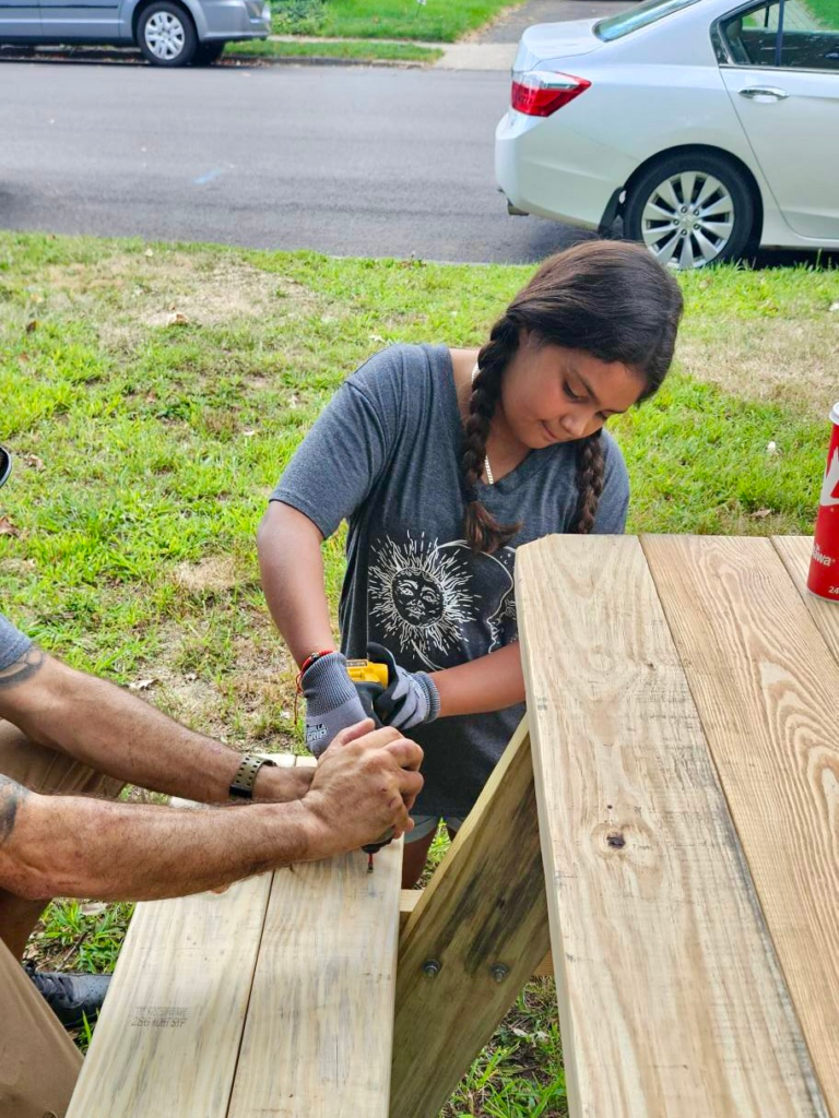 Girl Scout uses a drill on a picnic table plank while assisted by an adult volunteer
