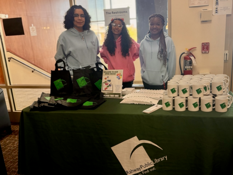 Three volunteers standing behind a table with Rahway Public Library tote bags and mugs at the library’s open house.