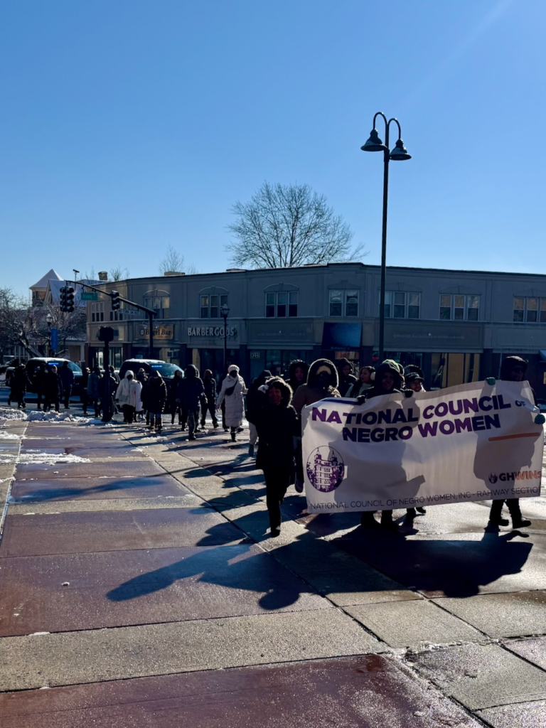 Community members march through downtown Rahway on Martin Luther King Jr. Day, carrying banners and walking together along snow-lined streets.