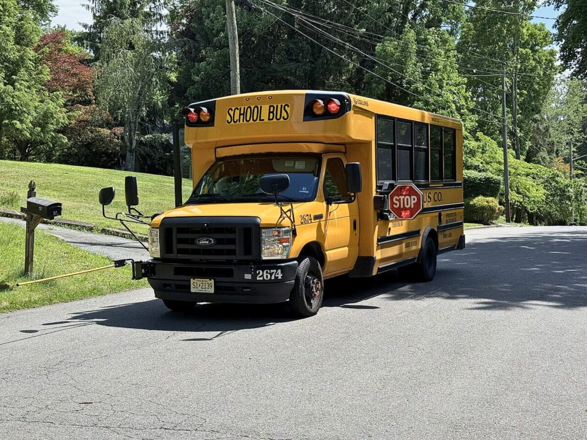 A small yellow school bus travels along a rural or suburban road in Mercer County, New Jersey, as part of a shared student transportation route.